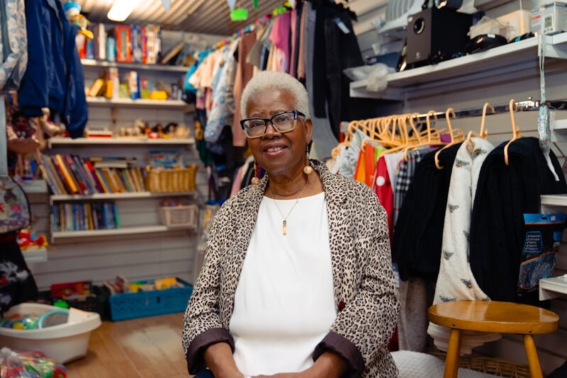 An older woman smiling in a community shop surrounded by clothes and books