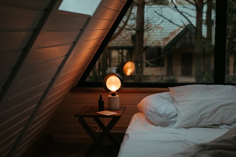 A bed beside a window at dusk with a warm lamp lit on the bedside table
