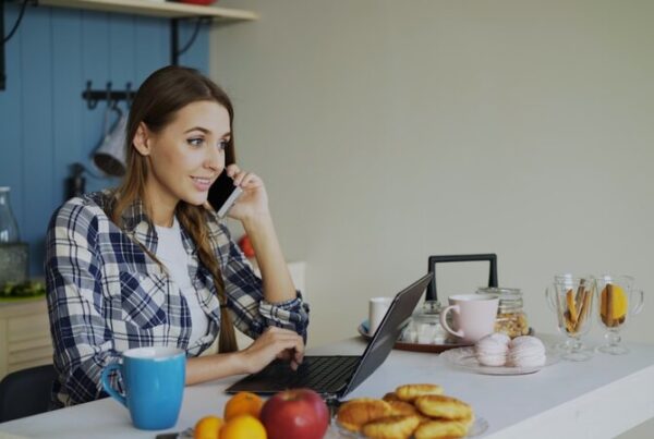 A woman sits at her kitchen table taking a phone call while working on a laptop, balancing work and caring responsibilities