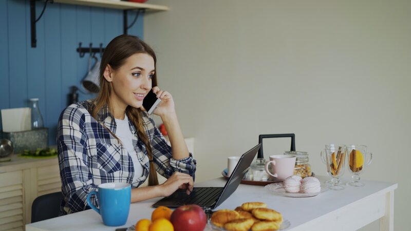 A woman sits at her kitchen table taking a phone call while working on a laptop, balancing work and caring responsibilities