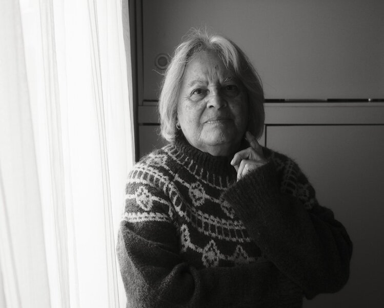 An older woman in a patterned jumper sitting quietly by a window in morning light